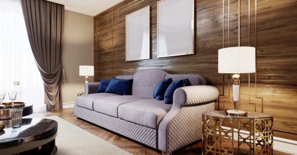 Luxurious soft sofa in the living room, purple and blue, against the background of a wooden wall with side gilded little tables on the side with table lamps, two empty paintings on the wall.